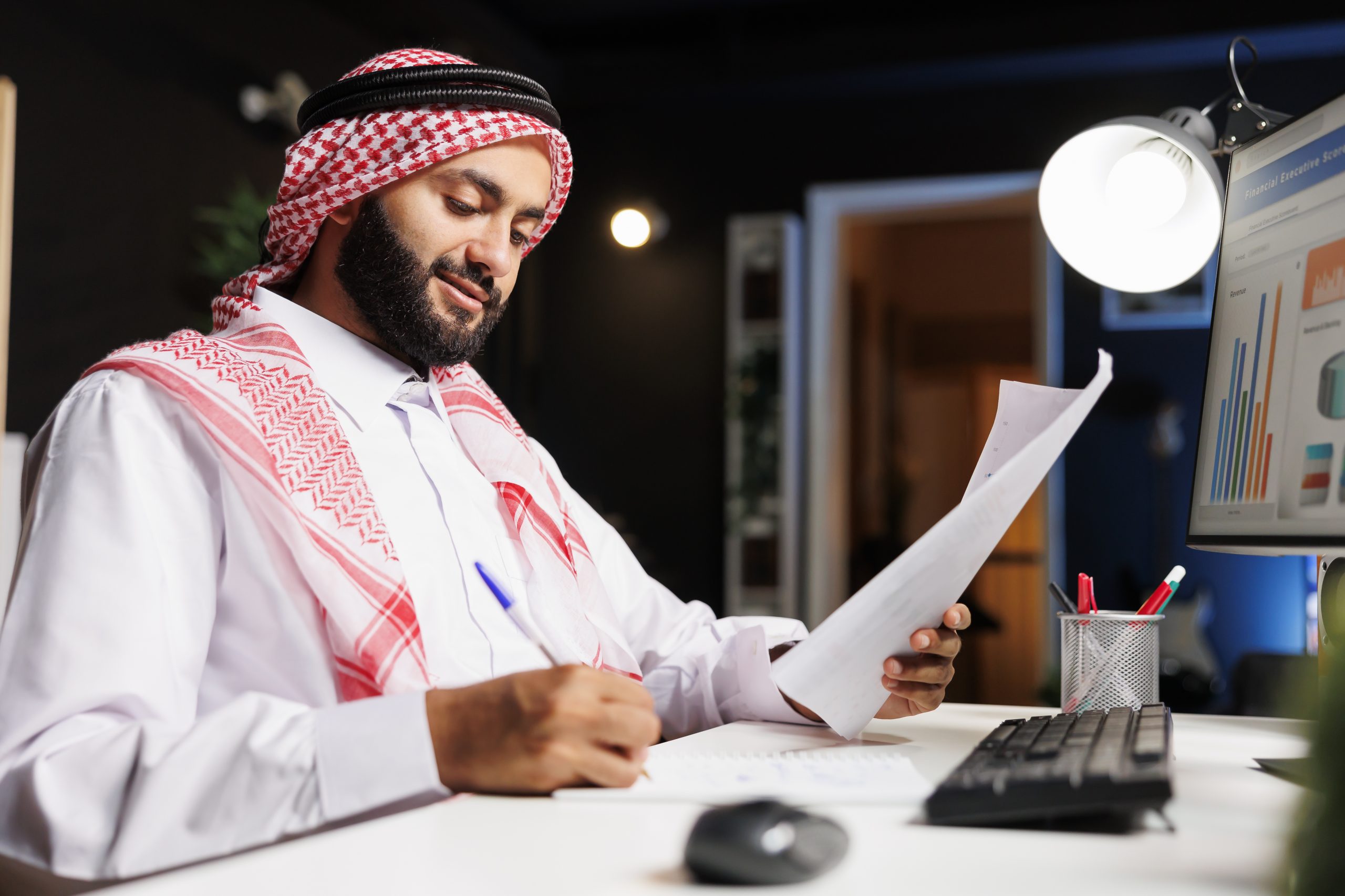 Islamic man working at his desk, surrounded by a desktop computer, documents, and paperwork, showcasing a busy corporate workspace. Side-view shot of Muslim guy writing down notes from research.